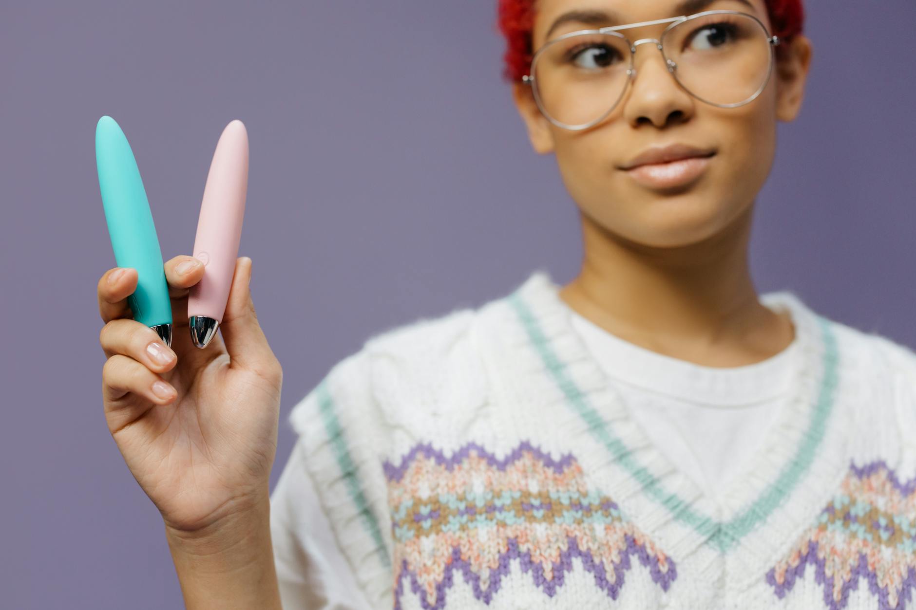 A woman holding two colorful vibrators against a purple background, representing pleasure and wellness tools.